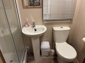 A bathroom with a white sink, toilet, and bidet. There is a glass shower enclosure, a toilet brush, and a trash bin.