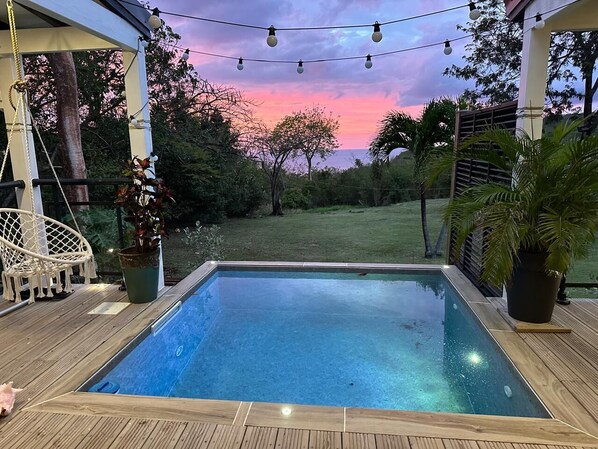 A rectangular swimming pool with a wooden deck surrounding it, under a pergola with string lights. - Gîte Avec Vues sur Mer, Piscine, Barbecue ,jardin (Deshaies)