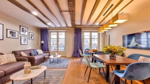 A modern living room with a sofa, coffee table, dining table, and chairs. There are framed pictures on the wall, a window with curtains, and a painting above the dining table.