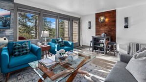 Ein modernes Wohnzimmer mit einem blauen Sofa, einem grauen Sofa und einem Glastisch. Eine Holzvertäfelung und ein großes Fenster mit Blick auf Bäume sind ebenfalls vorhanden.
