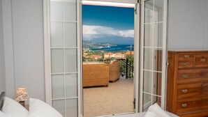 Un dormitorio con un amplio ventanal corredera que da a un balcón con un sofá de mimbre y vistas a un pueblo costero.