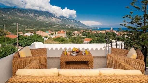 Una terraza con mobiliario de mimbre, una mesa con alimentos para el desayuno y una vista panorámica de un pueblo costero y montañas.