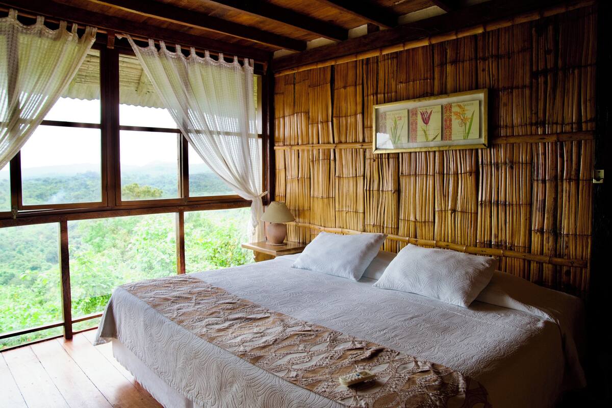 Quarto com uma cama grande, roupa de cama branca, cabeceira de madeira e vista para o verde através de janelas amplas.