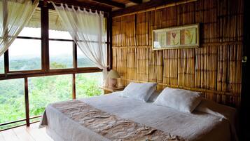 A bedroom with a large bed, white linens, a wooden headboard, and a view of greenery through large windows.