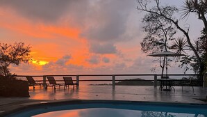A poolside area with lounge chairs and a table, overlooking a body of water with a sunset in the background.