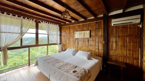 A room with a large bed, wooden ceiling beams, a window with a view of greenery, and an air conditioning unit.