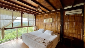 A room with a large bed, wooden ceiling beams, a window with a view of greenery, and an air conditioning unit. - Samai Lodge Holistic Living (Manglaralto)