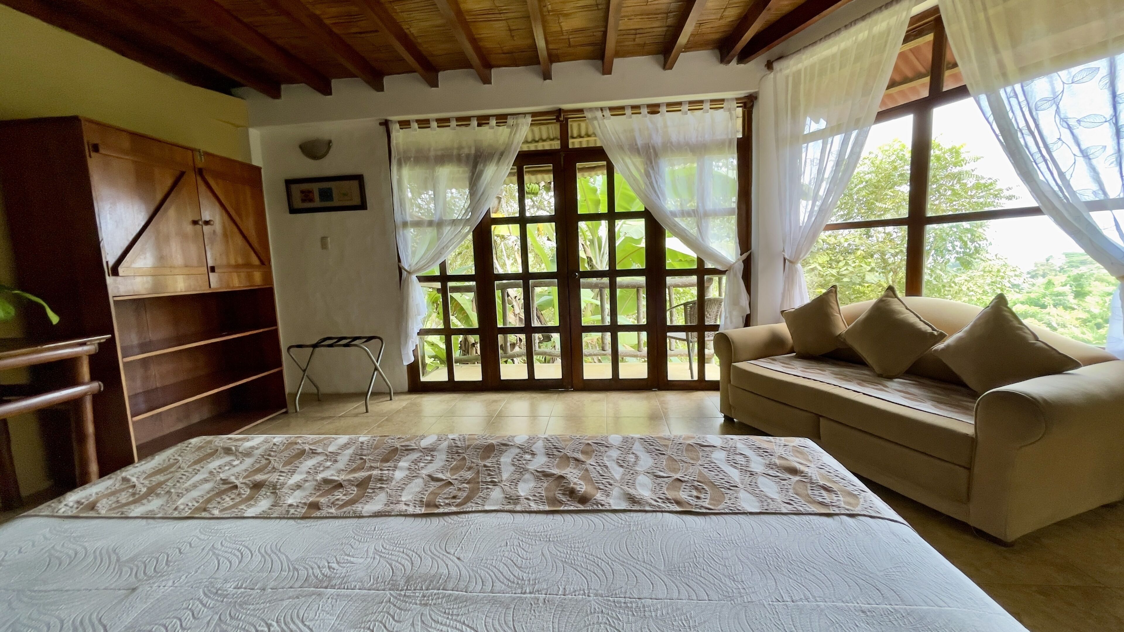 A bedroom with a bed, a sofa, a wooden wardrobe, and a window with curtains.