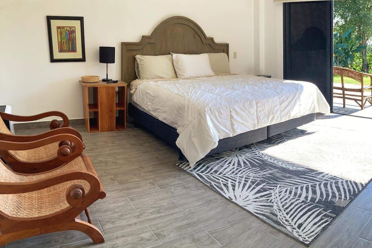 Um quarto com cama, mesas de cabeceira de madeira, uma cadeira de fibra natural, tapete estampado e um quadro na parede.