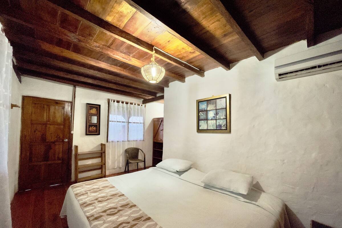 Quarto com teto de madeira, cama com roupa de cama estampada, uma cadeira, janela com cortinas e ar condicionado fixado na parede.