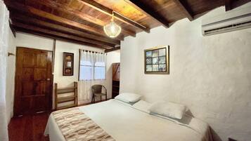 Un dormitorio con techo de madera, una cama con ropa de cama estampada, una silla, una ventana con cortinas y un aire acondicionado instalado en la pared.