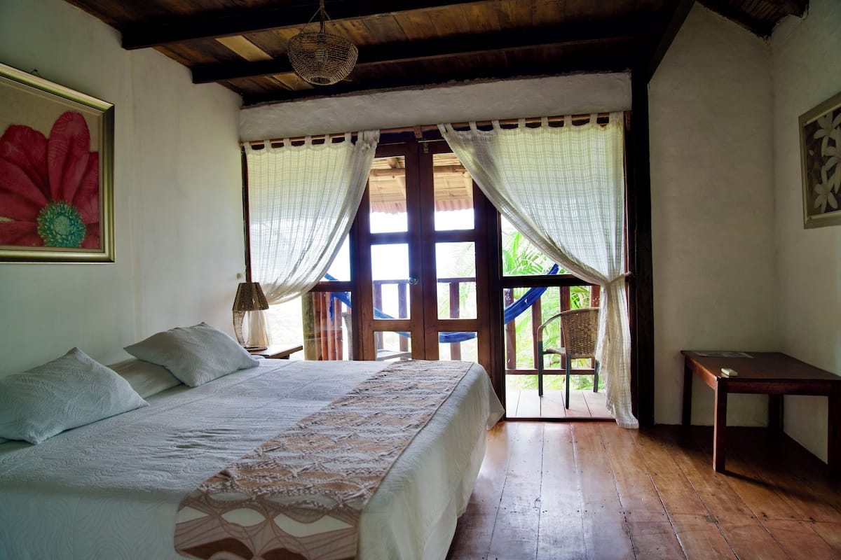 Quarto com cama, piso de madeira, uma mesinha e vista para o exterior.
