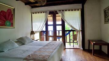 Un dormitorio con cama, piso de madera, una mesita y vistas al exterior.