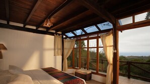 A bedroom with a large bed, a bench, and a view of the outdoors through a large window.