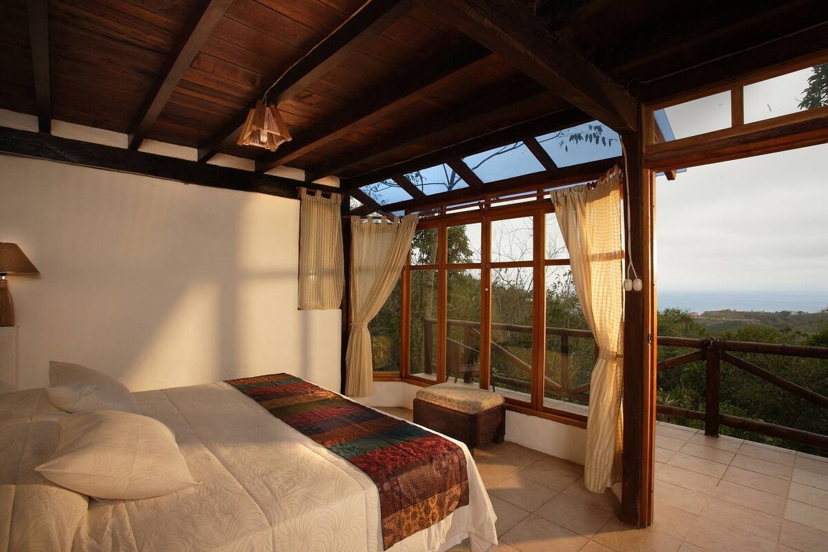 Quarto com uma cama grande, um banco e vista do exterior através de uma janela ampla.
