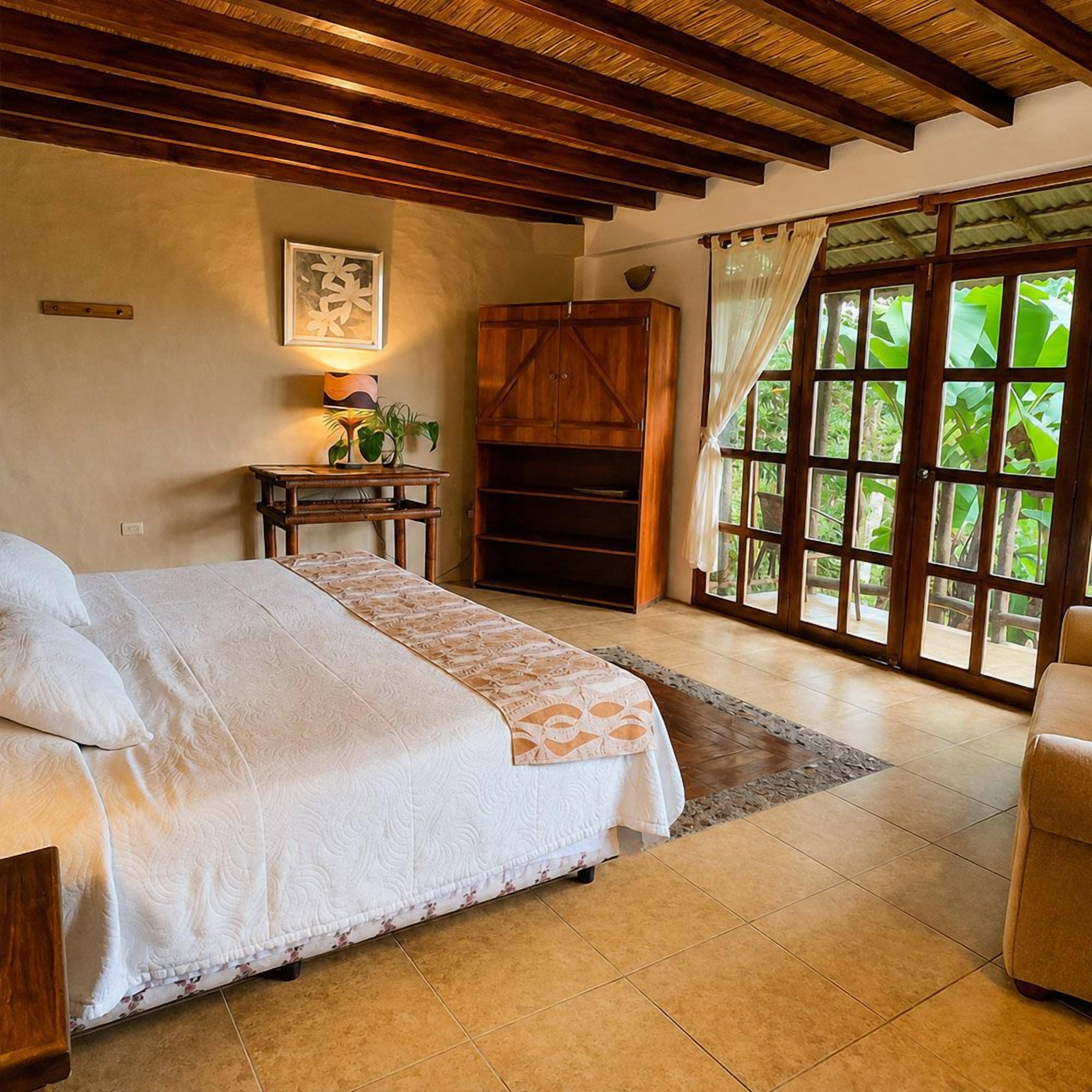 Un dormitorio con cama, armario de madera, mesita de noche con lámpara y vistas a través de un ventanal.