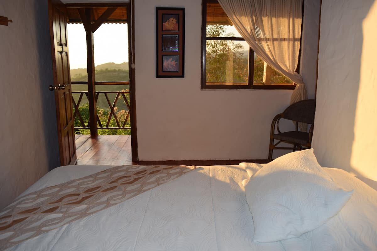 Quarto com cama, uma cadeira e varanda com vista para as colinas.