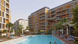 A swimming pool surrounded by lounge chairs and palm trees, with multi-story buildings in the background. - Exquisite 2BR w/ Assistant’s Room at Mesk 1 Midtown Dubai Production City by Deluxe Holiday Homes (Dubai)