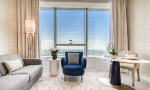 Ein modernes Hotelzimmer mit großem Fenster, einem grauen Sofa, einem blauen Sessel, einem runden Tisch mit weißer Schüssel und einem Blumentopf.