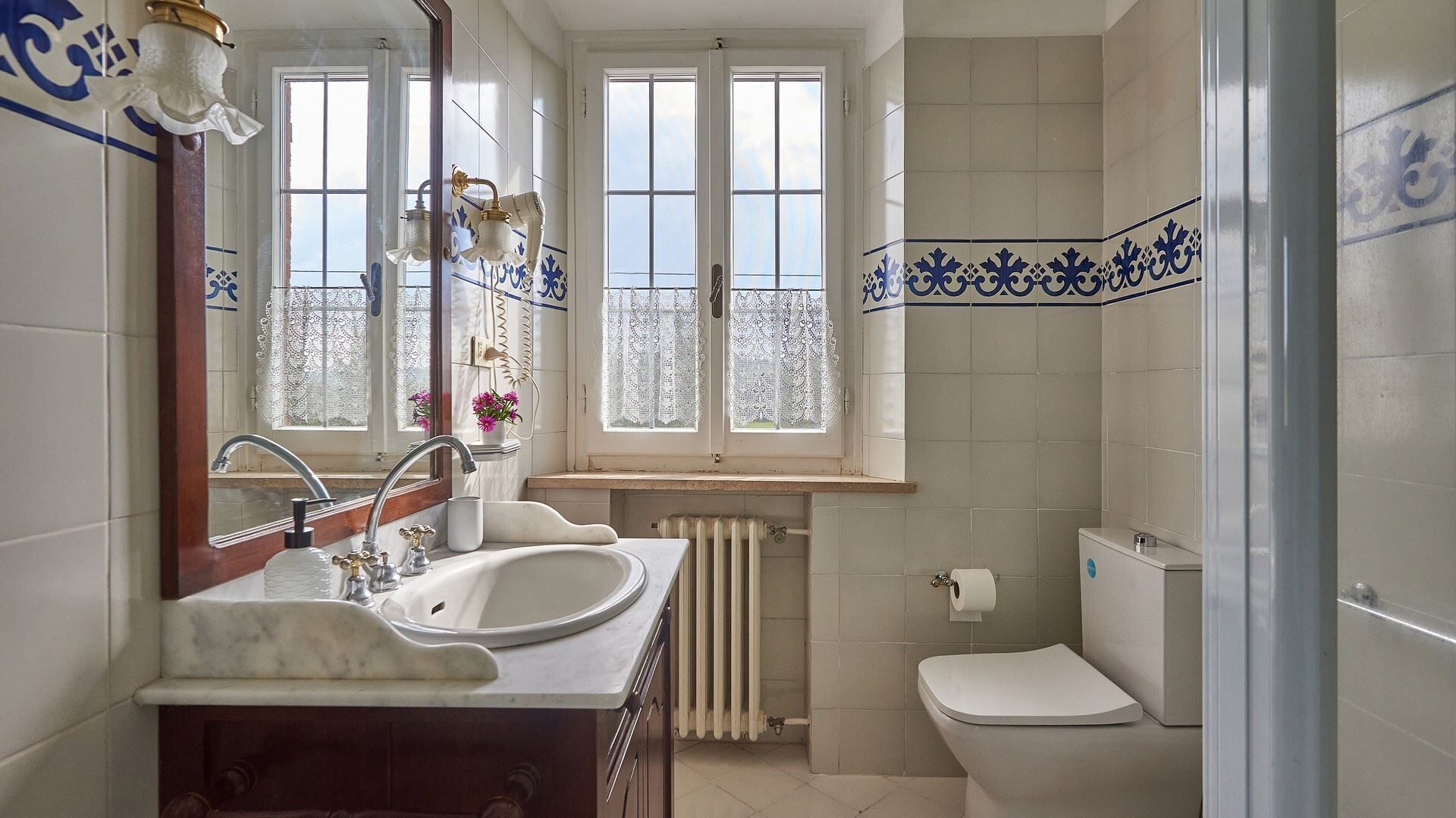 Een badkamer met een wastafel met twee kommen, een toilet, een radiator en een raam met kantgordijnen.