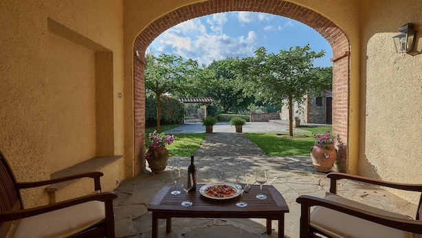 Una terrazza esterna con un tavolo apparecchiato per due persone, vista di un giardino attraverso un arco e un vialetto in pietra che conduce a un cancello.