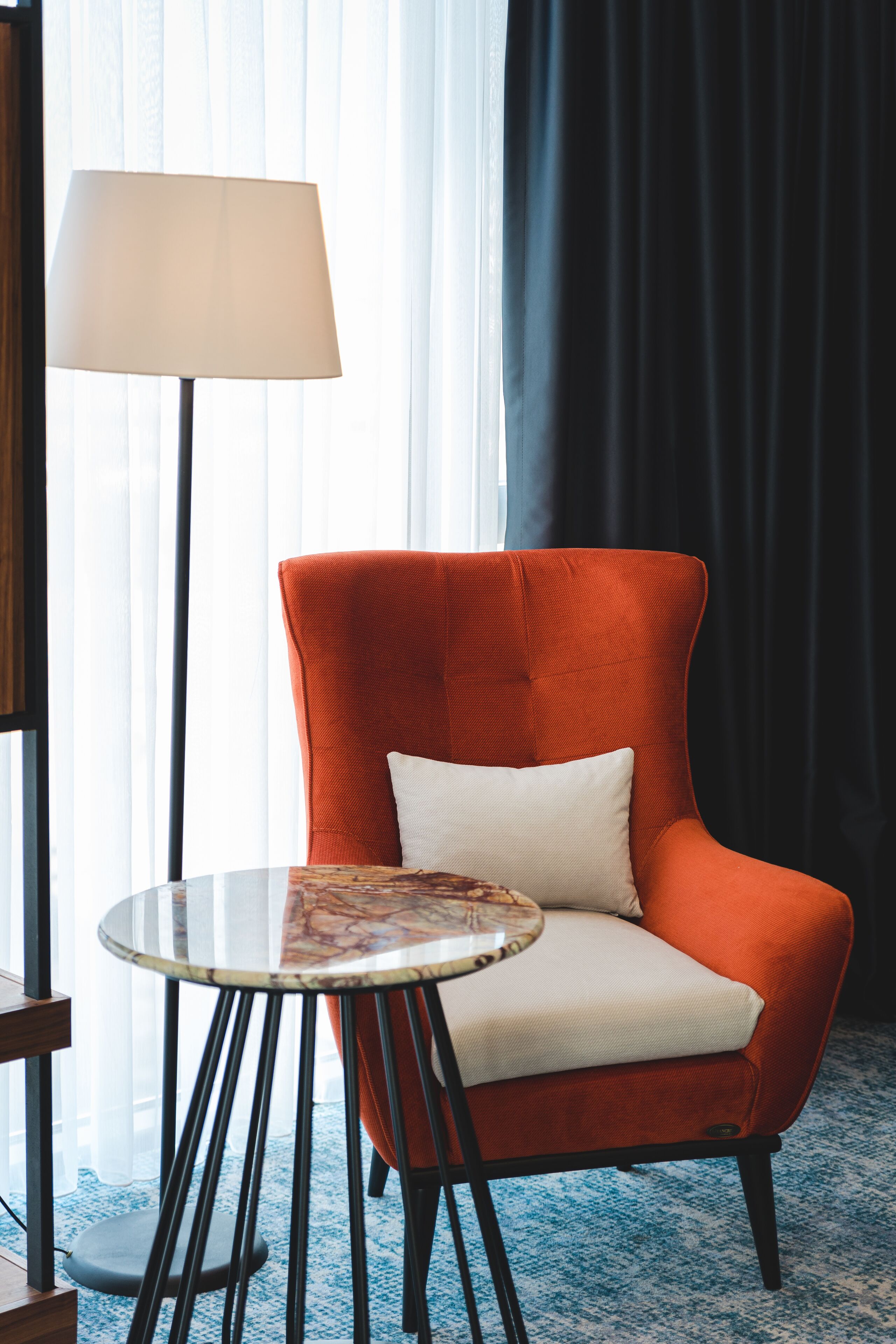 A room with a red armchair, a round table with a white cushion, a floor lamp, and a window with sheer curtains.