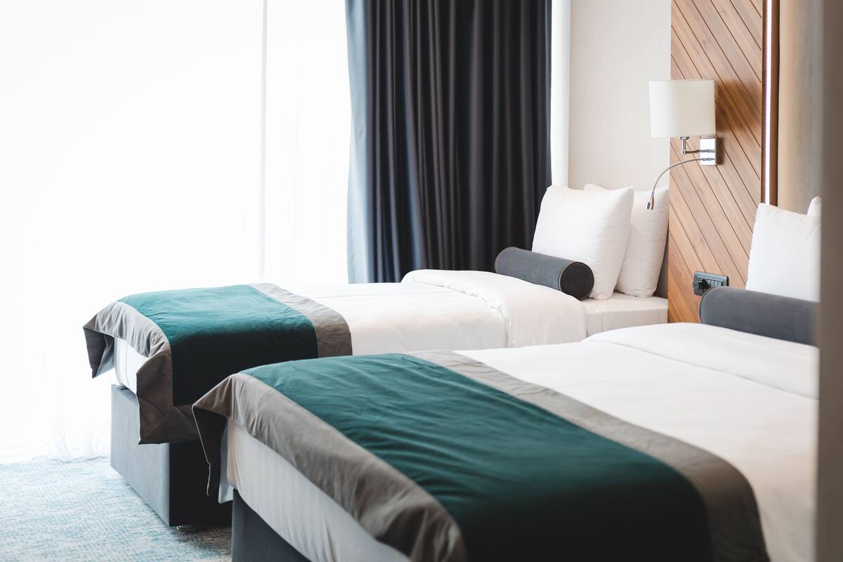 Habitación de hotel con dos camas, una mesita de noche, una lámpara y vistas al exterior.