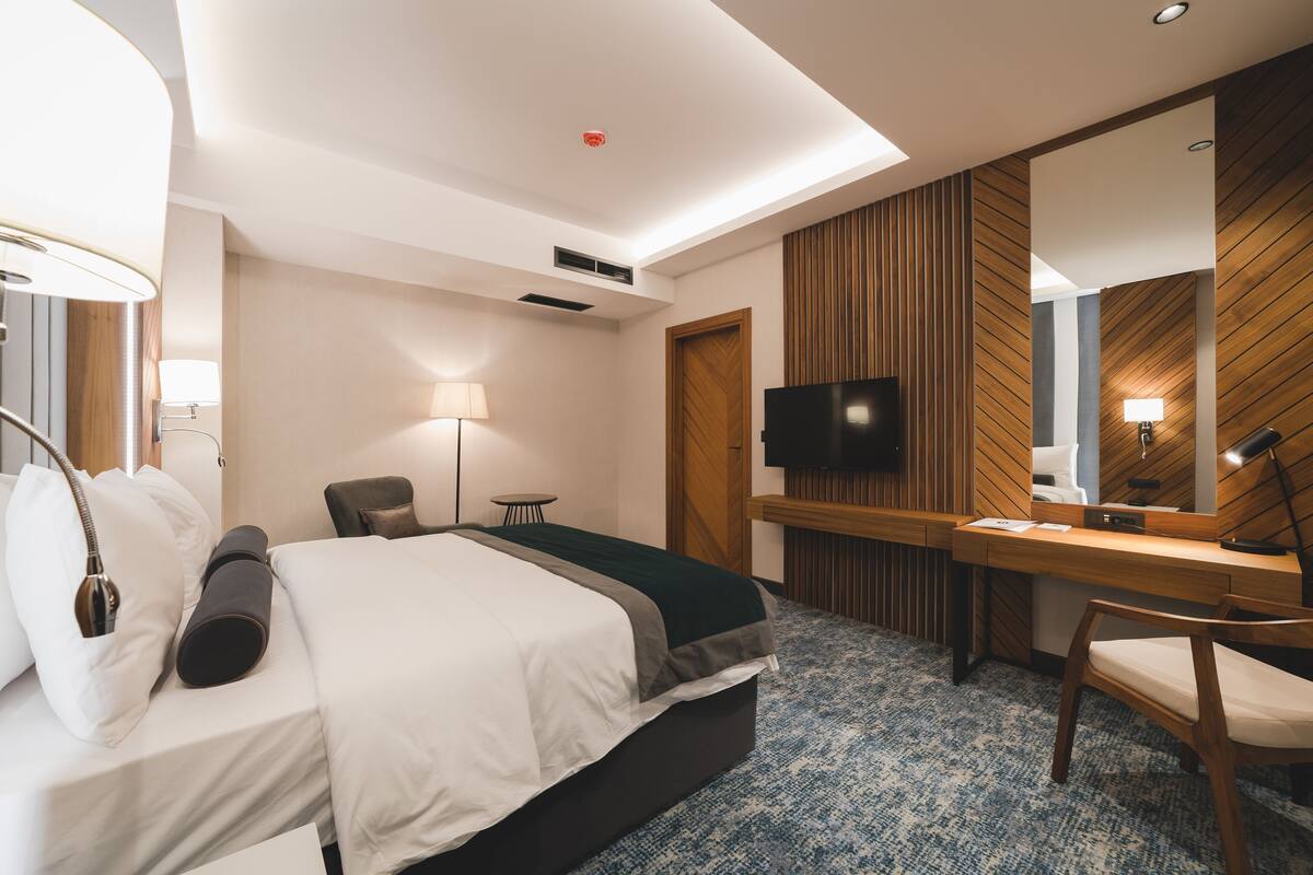 Una habitación de hotel moderna con cama, mesita de noche, lámpara, televisión y un escritorio con espejo.