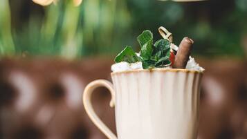 A cup of coffee with a biscuit and mint leaves on a saucer, with a blurred background of greenery and lights.