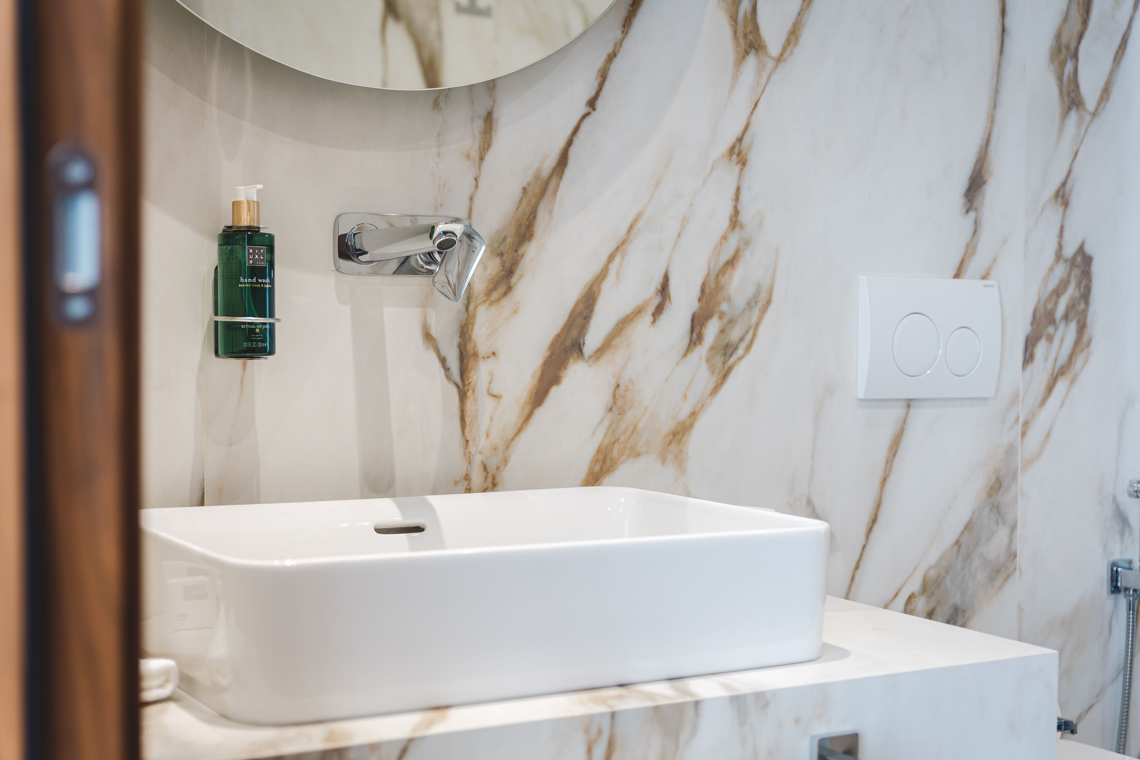 A bathroom with a white sink, marble walls, a mirror, and a hand soap dispenser.