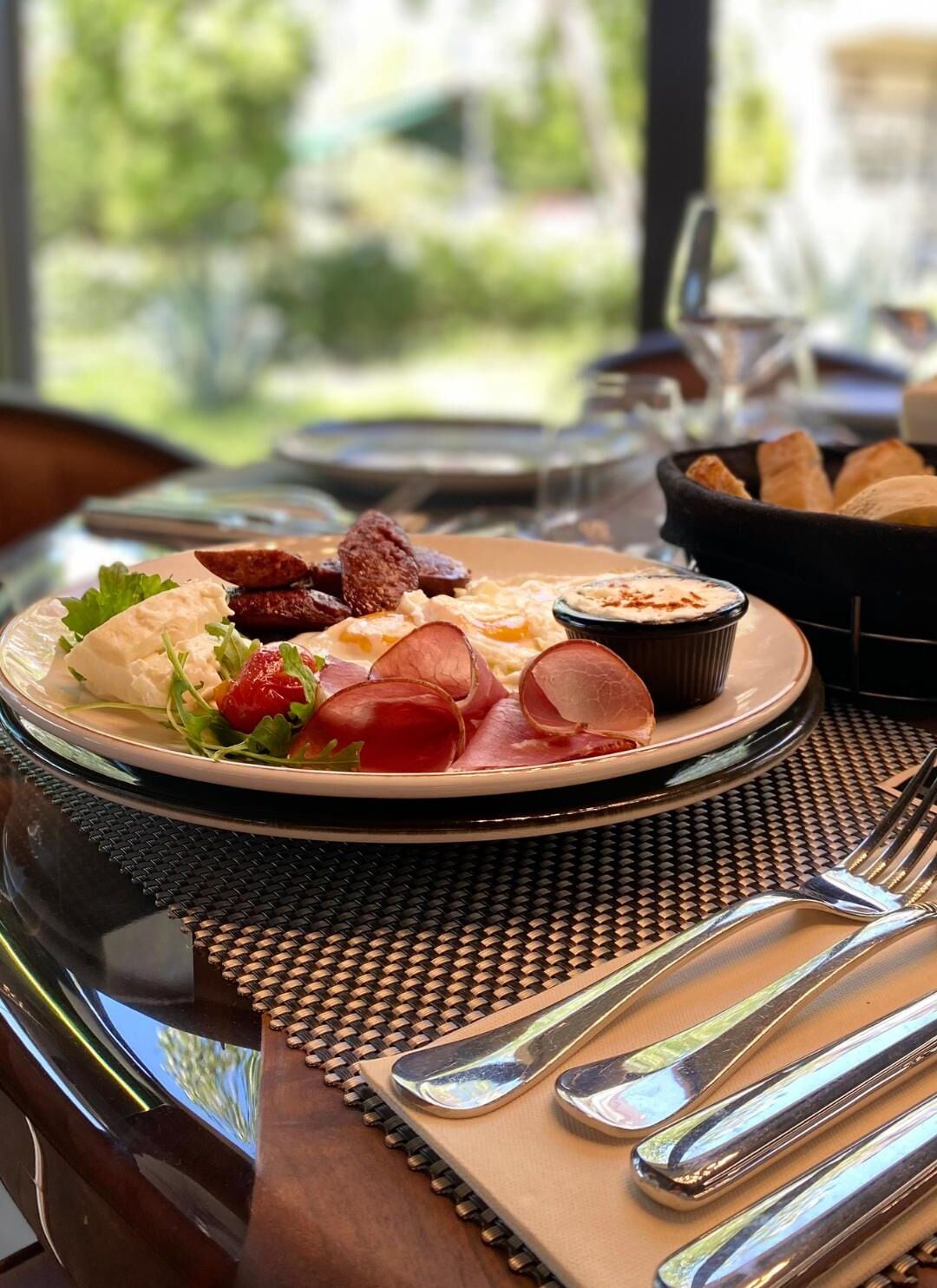 Un plato con una variedad de embutidos, quesos y un recipiente pequeño con salsa, sobre una mesa con un posavasos tejido y cubiertos.