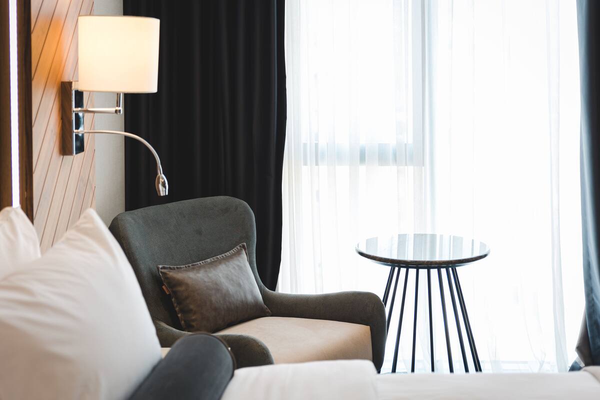 Habitación de hotel con cama, una silla, una mesita redonda y una lámpara de pared.