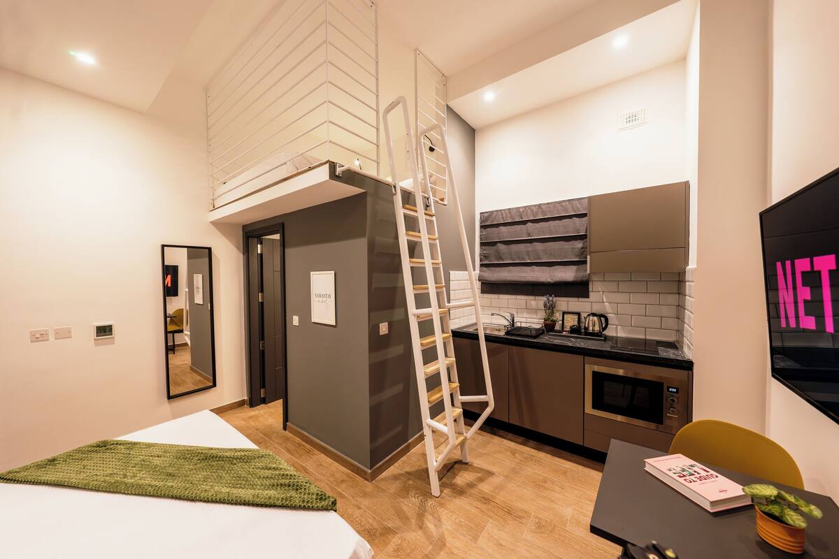 A modern apartment with a loft-style bedroom, a kitchen with a black countertop, and a dining area with a table and chair.