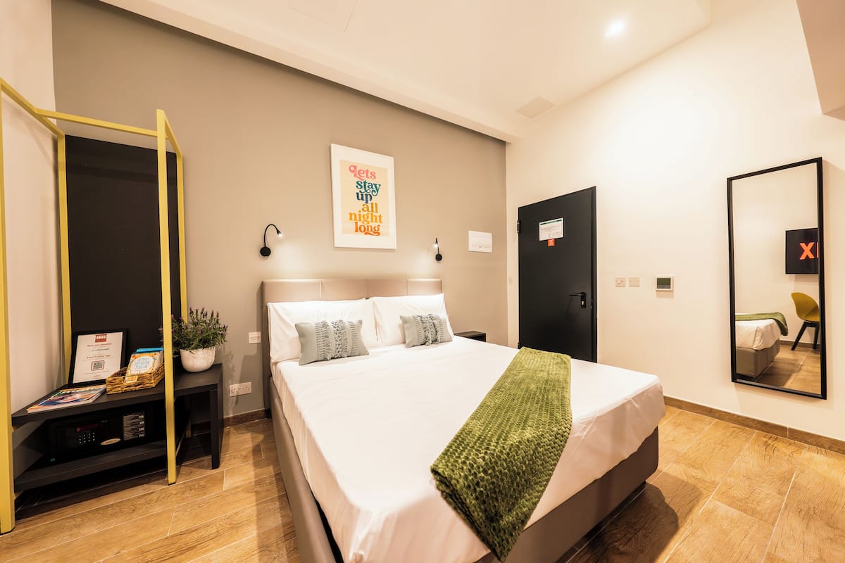 A modern hotel room with a large bed, a bedside table with a plant, a wall-mounted artwork, and a mirror reflecting the room.