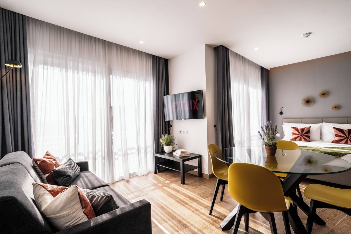 A modern living room with a sofa, a glass dining table, and yellow chairs. There is a TV mounted on the wall and a bed with pillows in the background.