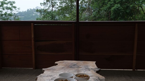 A dining table with bowls of food, plates, and cutlery set on a wooden table outdoors. - Camp David (Kohima)