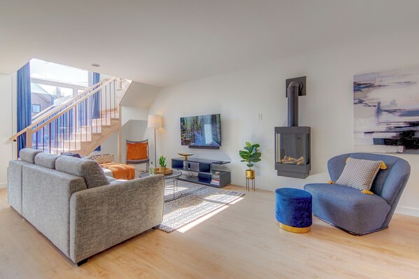 A modern living room with a sofa, armchair, TV, fireplace, and staircase.