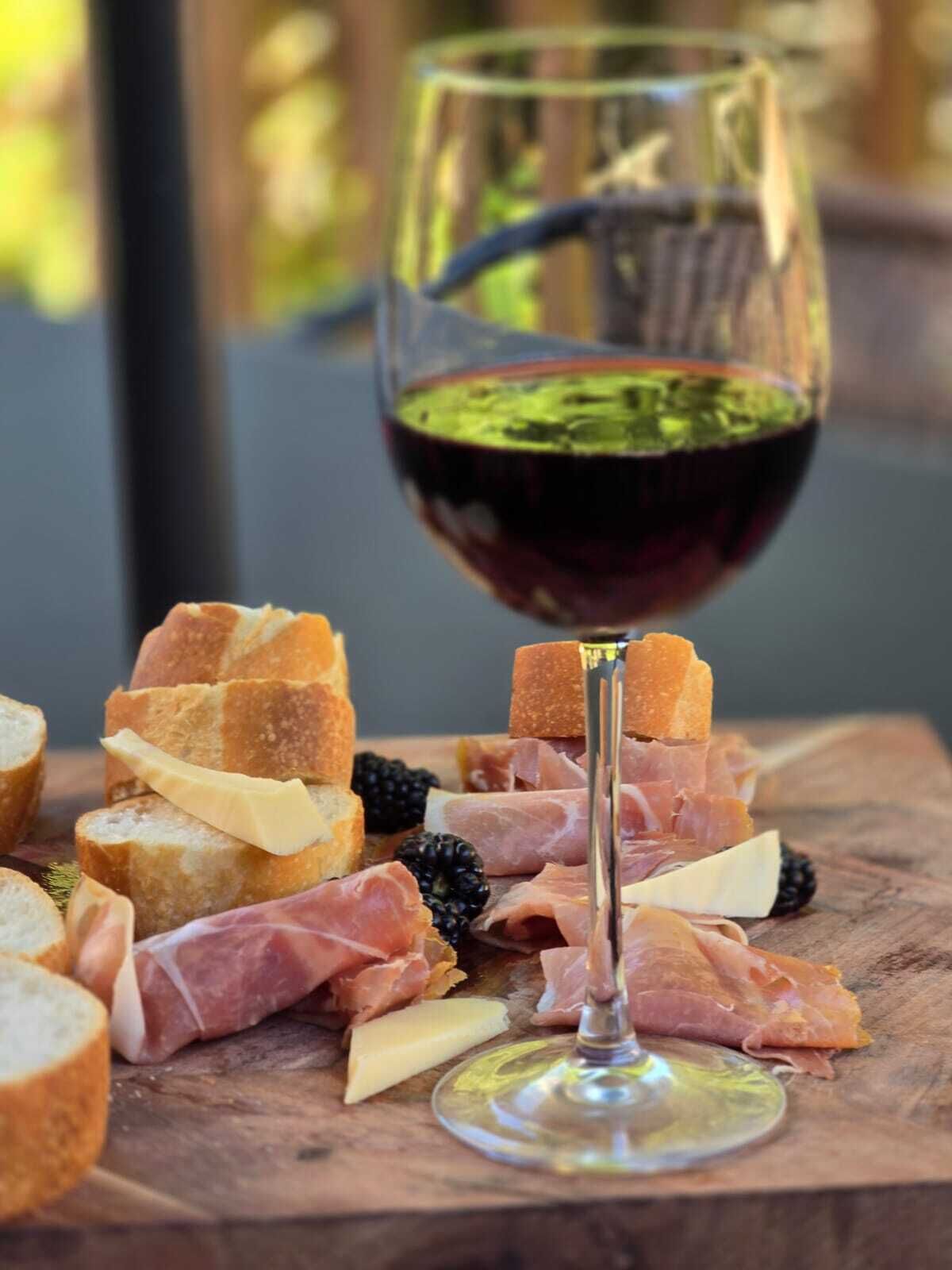 A glass of red wine with a green garnish, surrounded by various cheeses, cured meats, and bread on a wooden board.