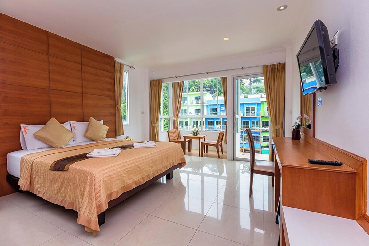 A hotel room with a bed, a desk, a TV, and a balcony with a view of buildings.
