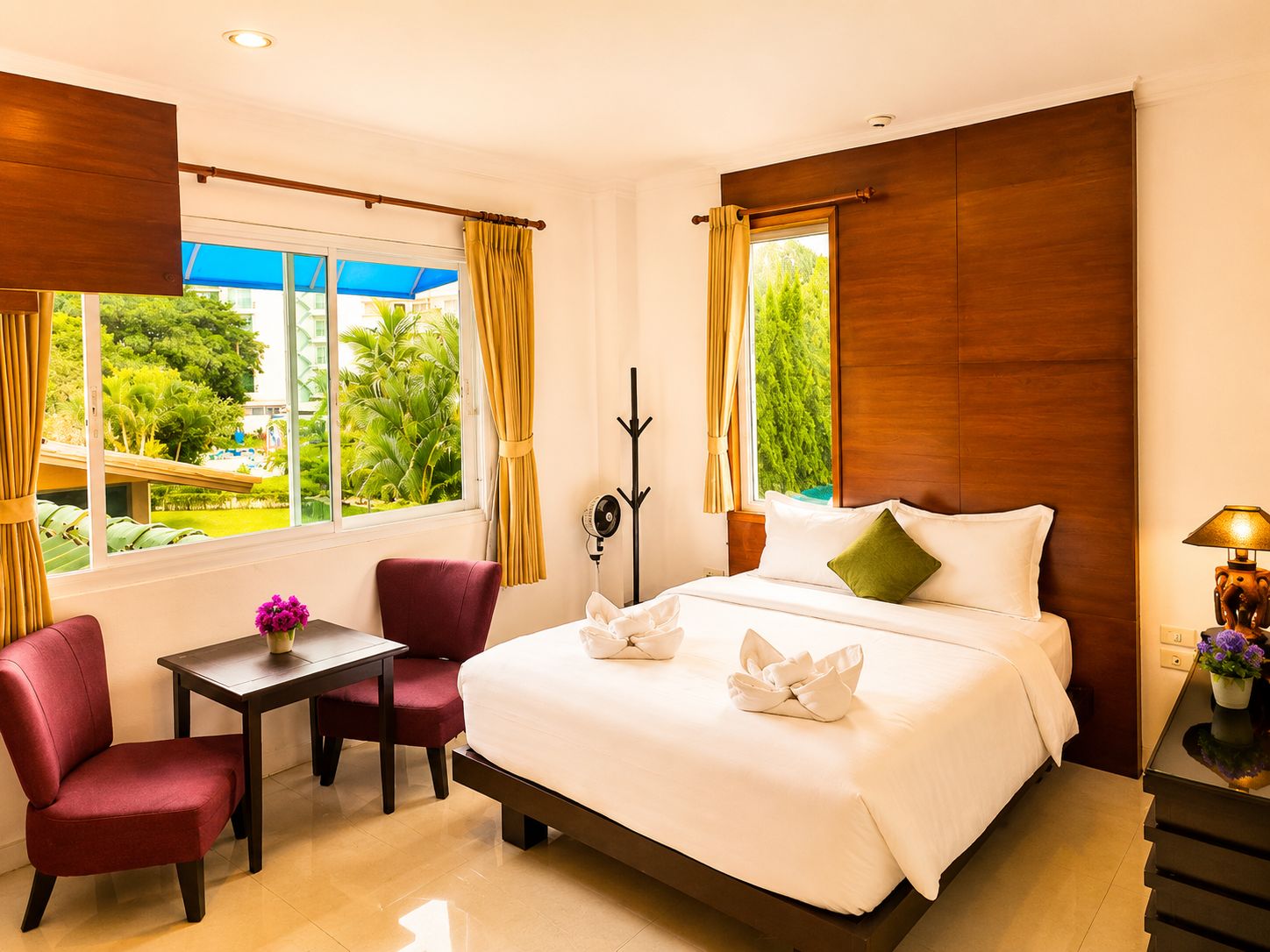 A hotel room with a bed, a desk, two chairs, a table with a vase of flowers, and a view of greenery outside.