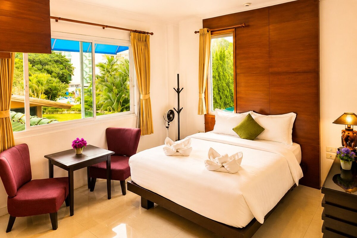 A hotel room with a bed, a desk, two chairs, a table with a vase of flowers, and a view of greenery outside.