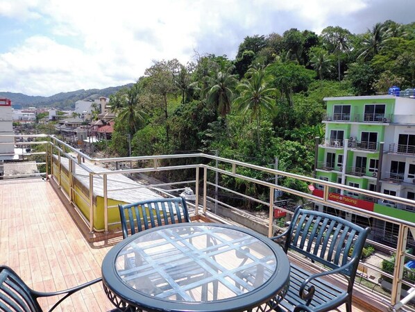 Une terrasse sur le toit, dotée d’une table et de chaises en verre, entourée d’une rambarde métallique, et offrant une vue sur un paysage urbain avec des immeubles et des palmiers.