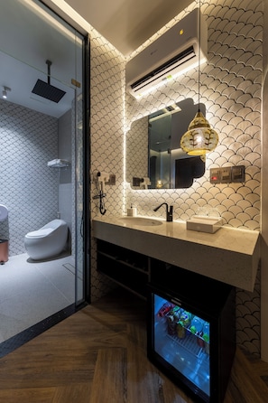 Un baño moderno con mampara de vidrio, inodoro, lavamanos con espejo, lámpara colgante y televisión integrada.