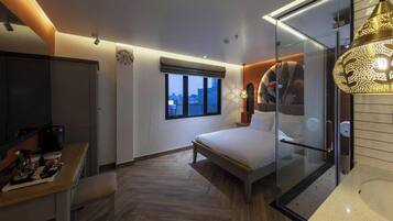 A modern hotel room with a bed, a desk with a coffee maker, a window, and a glass partition.