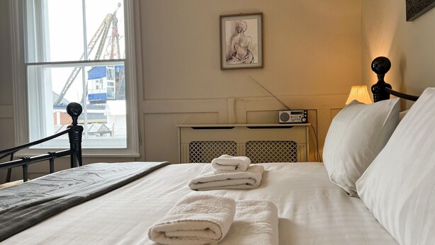 A neatly made bed with folded towels, a radio on a bedside table, and a framed artwork on the wall. - Bell Cottage (Cowes)