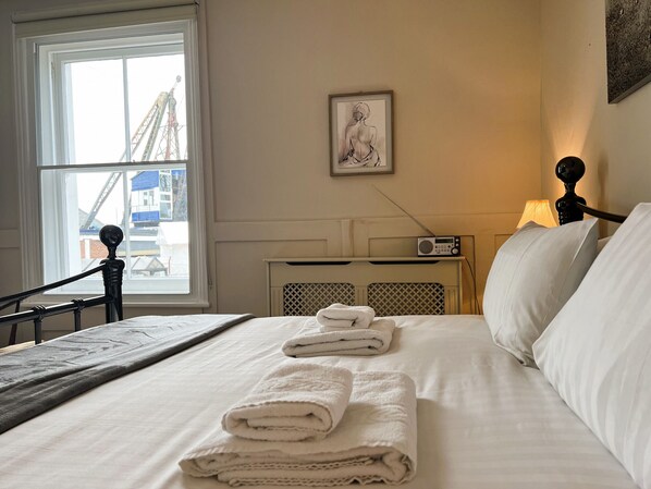 A neatly made bed with folded towels, a radio on a bedside table, and a framed artwork on the wall. - Bell Cottage (Cowes)