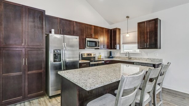 A modern kitchen with dark wood cabinets, stainless steel appliances, a granite countertop, and a central island with bar stools. - Downtown - Coastal Cozy 3B Home! (Pensacola)