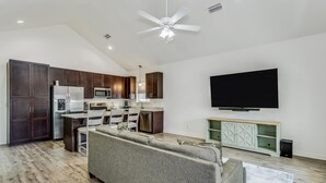 A modern kitchen with dark wooden cabinets, a stainless steel refrigerator, and a built-in microwave. There is a gray sofa in the living area, a flat-screen TV mounted on the wall, and a ceiling fan. - Downtown - Coastal Cozy 3B Home! (Pensacola)
