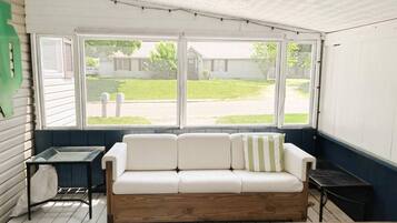 A room with a white sofa, a small table, and a large window with a view of a lawn and a house.