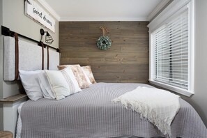 Un dormitorio con cama, almohadas, un edredón, una lámpara de pared, una corona decorativa y una ventana con persianas.